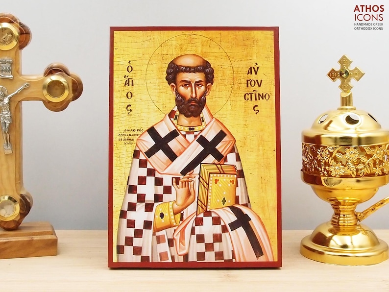 May include: An Orthodox icon featuring a saint with a halo, holding a book. The icon has a gold background and is framed in red. The saint is depicted in a checkered robe with black crosses. A gold censer and a wooden cross are on either side.