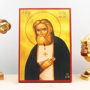 Może przedstawiać: Ręcznie malowana ikona Świętego Gerasimosa z Kefalonii, świętego prawosławnego. Ikona znajduje się na drewnianej tabliczce z czerwoną ramką. Święty jest przedstawiony z długą białą brodą i czarną szatą. Ikona jest otoczona złotą aureolą i złotym tłem.