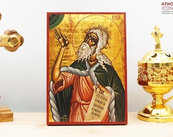 May include: Hand-painted icon of the prophet Elijah, with a red, gold, and green background. The icon is framed in a red wooden frame. The text on the scroll reads "ZH KYPPO O OEOE IS ANANMH O OEOE IS IEPIA".