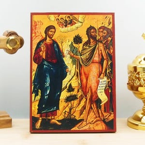 Puede incluir: Un icono pintado a mano que representa el bautismo de Jesucristo. El icono muestra a Jesús de pie en un río con Juan el Bautista vertiendo agua sobre su cabeza. El Espíritu Santo se representa como una paloma que desciende del cielo. El icono está pintado en un estilo bizantino tradicional con colores vibrantes y detalles dorados.