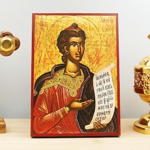 May include: A religious icon featuring a figure in red and black robes, holding a scroll with Greek text. The icon has a gold halo and is set against a gold background. A wooden cross and a gold censer are on either side.