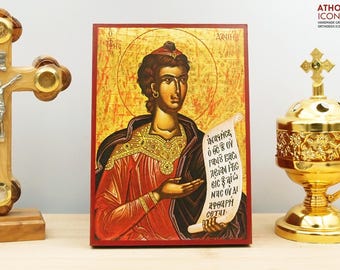 May include: A religious icon featuring a figure in red and black robes, holding a scroll with Greek text. The icon has a gold halo and is set against a gold background. A wooden cross and a gold censer are on either side.