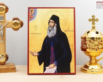May include: An Orthodox icon featuring a religious figure with a long white beard, wearing a black robe and hat. The icon has gold accents and Greek text. A gold incense burner and wooden cross are also visible.