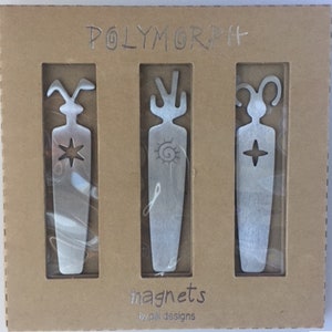 Puede incluir: Un juego de tres imanes de metal plateado en una caja de cartón. Cada imán tiene un diseño abstracto único, que incluye una estrella, un sol y una cruz. La caja está etiquetada como "POLYMORPH" y "magnets by pill designs."