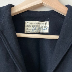 Könnte beinhalten: Eine schwarze Wolljacke mit einem Etikett mit der Aufschrift "Manufactured by Naval Clothing Factory". Das Etikett enthält auch einen handschriftlichen Namen und Rang: "J.C. Stone, R.M. 3/10".