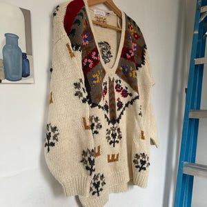May include: A cream-colored knitted sweater with a V-neck and a colorful geometric pattern. The pattern features flowers, leaves, and geometric shapes in shades of red, green, brown, yellow, and blue.