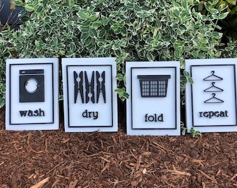 May include: Four white square signs with black borders and black icons and text. The signs read "wash", "dry", "fold", and "repeat".