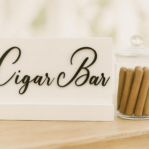 Puede incluir: Un letrero blanco con las palabras "Cigar Bar" en elegante escritura negra. Un tarro de cristal lleno de puros está junto al letrero. Flores blancas en el fondo. La escena está ambientada sobre una superficie de madera clara.