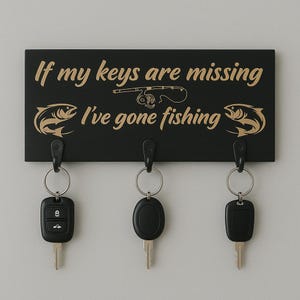 Può includere: Portachiavi nero con testo dorato "If my keys are missing I've gone fishing". Presenta una grafica di canna da pesca e tre ganci che tengono dei mazzi di chiavi. Il portachiavi ha una grafica di pesce.