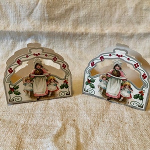 May include: Two vintage paper Christmas napkin holders featuring a girl holding a pie and two children. The holders are white with red and green accents and a holly design.