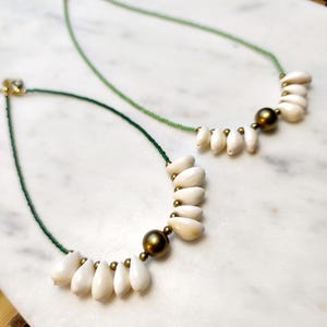 May include: Two necklaces with white shell pendants and green beaded cords. Each necklace features a cluster of white shell pendants, a large gold-colored bead, and smaller gold-colored beads. The necklaces are displayed on a white marble surface.