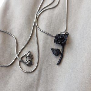 May include: A silver-colored necklace with a black rose pendant. The rose pendant has detailed petals and a stem with a single leaf. The necklace chain is a thin, snake-style design, and the clasp is visible.