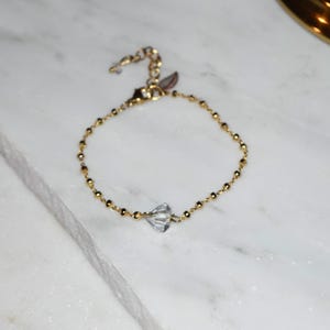 Puede incluir: Una delicada pulsera dorada con pequeñas cuentas redondas y un dije central de cristal transparente. La pulsera tiene una cadena ajustable y un pequeño dije de hoja cerca del cierre. La joya se exhibe sobre una superficie de mármol blanco.