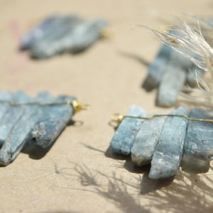 May include: A set of five blue kyanite crystal pendants, each wrapped with gold wire, arranged on a light brown surface.