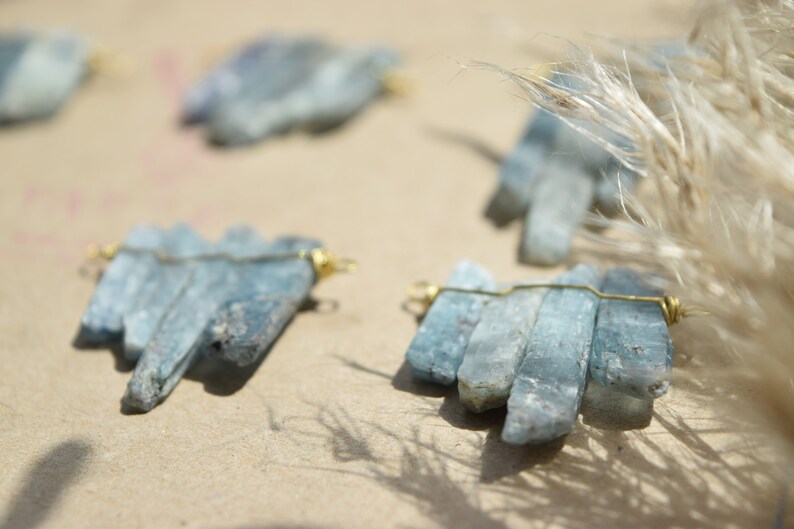 May include: A set of five blue kyanite crystal pendants, each wrapped with gold wire, arranged on a light brown surface.