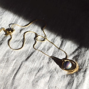 May include: A gold-colored necklace with a pendant featuring a round, iridescent stone. The necklace is lying on a white, textured surface, with sunlight casting shadows across the fabric.