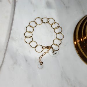 Pode incluir: Pulseira dourada com uma série de círculos texturizados interligados. A pulseira inclui um pequeno pingente com a letra "E". A joia é exibida em uma superfície de mármore branco.
