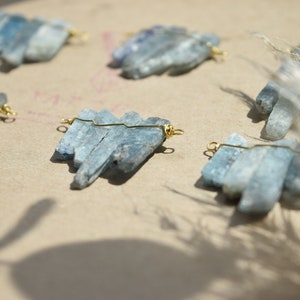 May include: Five sets of blue kyanite crystal clusters, each wired with gold jump rings, arranged on a light brown surface.