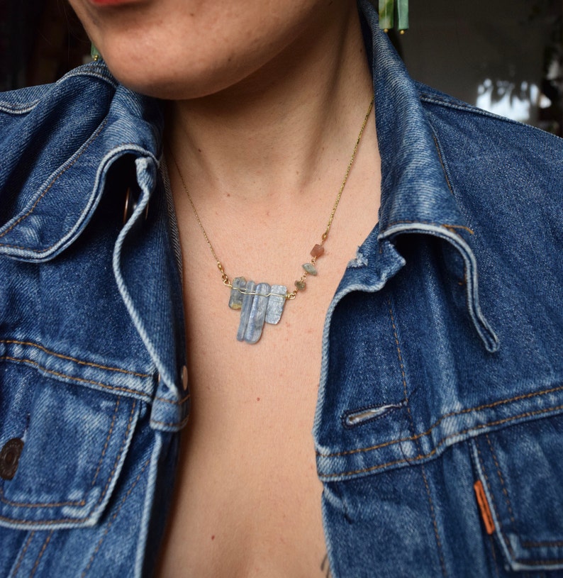 May include: A gold chain necklace with a cluster of blue-grey and brown crystal stones. The necklace is worn by a person wearing a denim jacket.