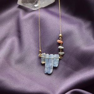 May include: A gold chain necklace with a pendant made of blue kyanite stones and small orange and brown stones. The necklace is on a purple fabric background.