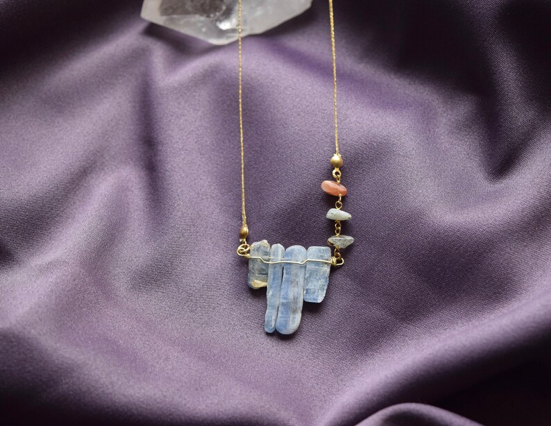 May include: A gold chain necklace with a pendant made of blue kyanite stones and small orange and brown stones. The necklace is on a purple fabric background.