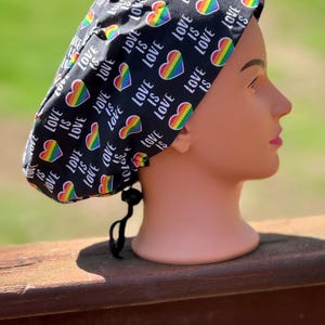 May include: Black scrub cap with a repeating pattern of rainbow hearts and the words "LOVE IS LOVE" in white. The cap is tied at the back with a black drawstring. The cap is displayed on a mannequin head.