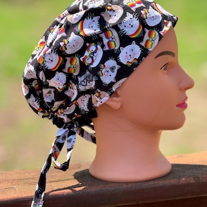May include: Black surgical scrub cap featuring a repeating pattern of cartoon cats with rainbow accents. The cap has a tie closure at the back. The cats have various expressions and are set against a black background.