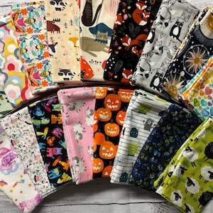 May include: A collection of 20 colorful fabric squares with various patterns, including animals, flowers, pumpkins, and stars. The fabric squares are arranged in a semi-circle on a white wooden surface.