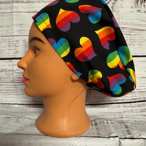 May include: A black scrub hat with a rainbow heart pattern. The hat is worn on a mannequin head.