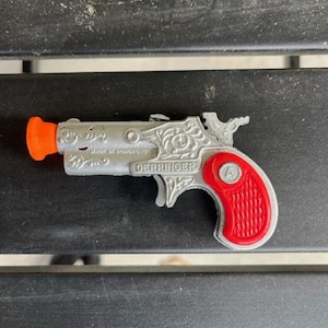 May include: A silver toy derringer with a red plastic handle. The toy gun has an orange plastic tip and the words "MADE IN HONG KONG" and "DERRINGER" engraved on the side.
