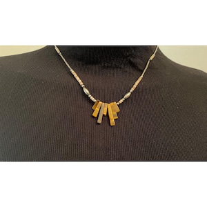 Puede incluir: Un collar con una cadena plateada y un colgante con piedras rectangulares de ojo de tigre en tonos marrón y dorado. El collar tiene pequeñas cuentas redondas y se muestra sobre un fondo negro.
