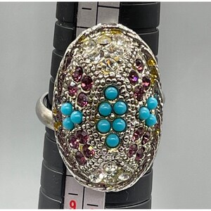 May include: Silver-toned ring with an oval-shaped face. The face is encrusted with clear, purple, and turquoise-colored stones. The ring is shown on a black ring sizer.