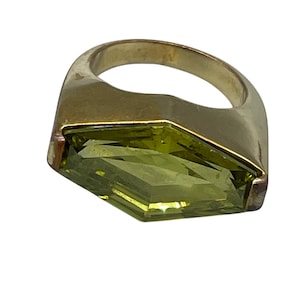 May include: A gold-toned ring featuring a large, faceted, light green gemstone. The gemstone is rectangular with angled sides, set in a thick, rounded band. The ring has a vintage or antique appearance.