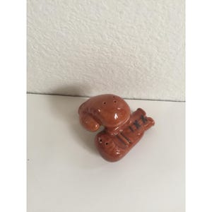 Puede incluir: Un juego de salero y pimentero de cerámica marrón. El salero tiene forma de guante de boxeo y el pimentero tiene forma de guante de boxeo. El juego está sobre una superficie blanca.