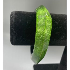 Könnte beinhalten: Ein leuchtend lindgrüner Armreif aus durchscheinendem Harz mit eingebetteten feinen Glitzerpartikeln. Der Armreif hat eine facettierte, geometrische Außenfläche mit mehreren kantigen Kanten und ein glattes, abgerundetes Innenband.