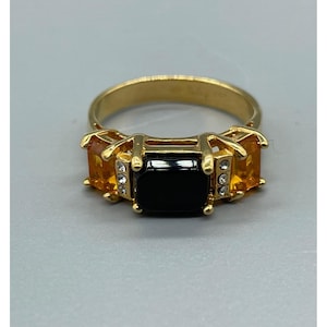 May include: Gold-toned ring featuring a large, rectangular black stone flanked by two smaller, orange-yellow stones. The ring has a classic design with small, clear accent stones. A statement piece of jewelry.