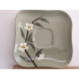 Puede incluir: Un platillo cuadrado de cerámica verde pálido con esquinas redondeadas. Presenta un diseño floral pintado de flores blancas, hojas verdes y un tallo marrón. El platillo tiene un centro empotrado para una taza de té.