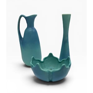 May include: A set of three ceramic objects in shades of blue and teal. The collection includes a tall pitcher with a curved handle, a slender vase with a narrow neck, and a bowl shaped like a flower with a perforated base for floral arrangements.