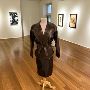 May include: A brown leather skirt suit displayed on a mannequin in an art gallery. The suit includes a long-sleeved jacket with a wrap-style closure and a matching knee-length skirt. The jacket is cinched at the waist with a belt.