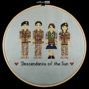 May include: Cross-stitch embroidery in a circular wooden frame depicting four figures in military uniforms. The figures are standing in a row, with the text "Descendants of the Sun" below them.