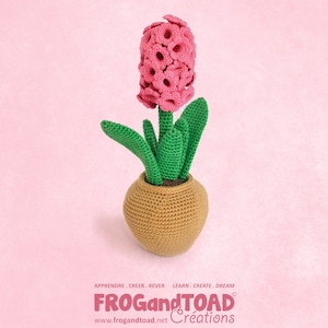 Puede incluir: Una flor de ganchillo hecha a mano en una maceta. La flor es de color rosa vibrante, con hojas y tallo verdes, y se encuentra en una maceta de color beige. El fondo es de color rosa suave. La base de la imagen tiene el texto "FROG and TOAD Creations".