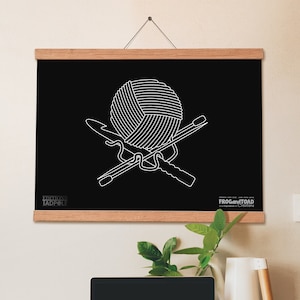 Op de afbeelding: Zwarte wandkunst met een witte illustratie van een bol wol en gekruiste haak- en breinaalden. Het kunstwerk wordt weergegeven in een houten frame en hangt aan een muur. Het kunstwerk is voorzien van de tekst "FROG and TOAD Editions TADPOLE".