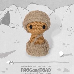 May include: A handmade crochet doll depicting a caveman, wearing a brown hat and fur-like clothing. The doll has black button eyes and a strap across its shoulder. The background features a cave drawing and the text "FROG and TOAD Créations".