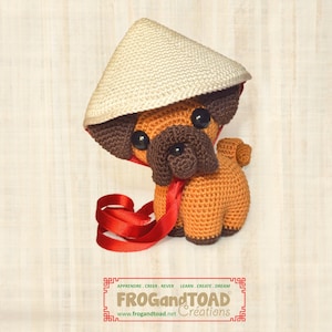 Può includere: Un carlino all'uncinetto con un cappello conico beige e un nastro rosso. Il carlino è marrone con dettagli marrone scuro e grandi occhi neri. L'immagine include il testo "FROG and TOAD Creations".
