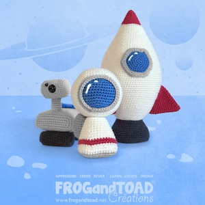 May include: Crocheted space-themed toys, including a rocket ship with red accents, a robot, and an astronaut figure. The toys are white, gray, and blue, with a blue background and the text "FROG and TOAD Creations".