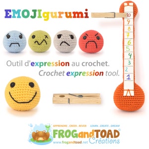 May include: A collection of crocheted emoji balls in various expressions, including sad, confused, angry, and happy. The image also features a wooden clothespin and a crocheted orange thermometer with numbers 1-10. Text reads "EMOJigurumi" and "Crochet expression tool."