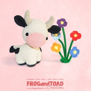 Könnte beinhalten: Eine gehäkelte Kuh mit schwarzen und weißen Flecken, Ohren und Hörnern sowie einer rosa Nase. Neben der Kuh befinden sich bunte gehäkelte Blumen mit grünen Stielen. Der Text "FROG and TOAD Créations" befindet sich unten.