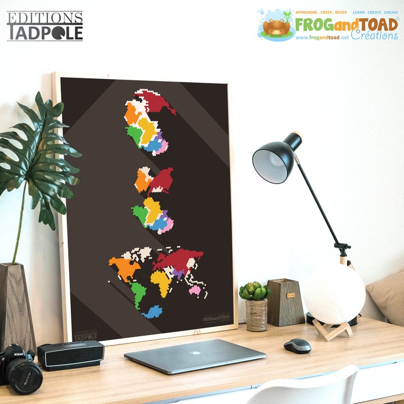 May include: A framed print featuring a pixelated world map in vibrant colors against a dark background. The print includes the text "EDITIONS TADPOLE" and "FROG and TOAD Creations". The artwork is displayed on a wooden desk with a laptop, lamp, and decorative items.