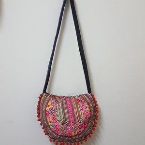 May include: A red, white and pink embroidered fabric bag with a black strap and red pom-poms around the edge.