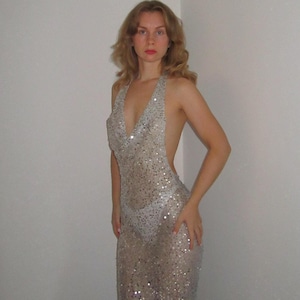 May include: A silver sequined halter dress with a low back. The dress is floor-length and has a cowl neckline. The sequins catch the light, creating a shimmering effect. The dress is form-fitting and sleeveless.